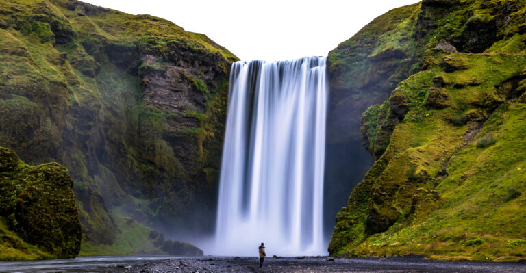 a lone male traveler experiencing the breathtaking and powerful Skogafoss waterfall in Iceland. scale of how huge the waterfall compared to the size of a man. By Nathaniel Gonzales